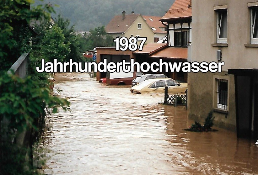 '1987 Jahrhunderthochwasser' text over a flooded German town street with cars partially submerged and water reaching doorways.